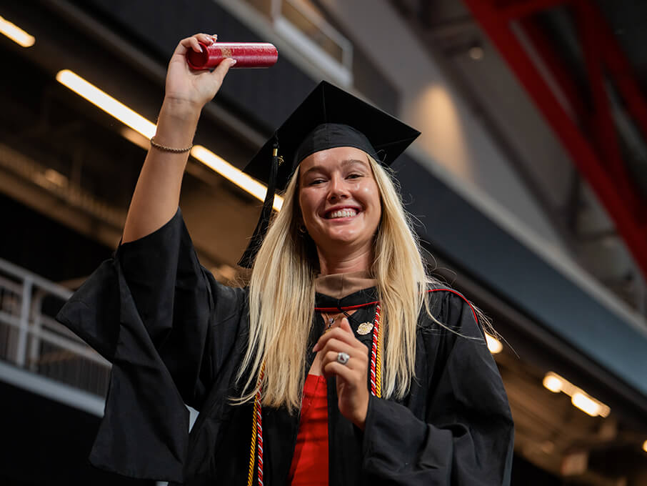 A woman in a graduation gown proudly holds up a red diploma case, celebrating her academic achievement.